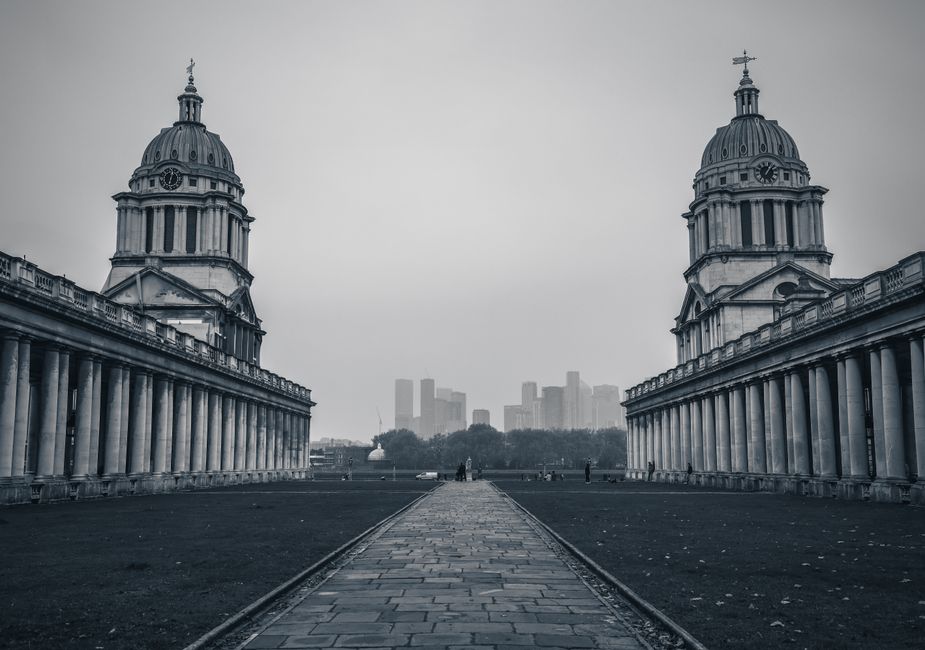 A black and white photograph of a spacious outdoor area between two symmetrical historic domed buildings with classical architectural features, including columns and balustrades. The foreground shows a paved pathway made of rectangular stone slabs leading towards a distant city skyline with modern high-rise buildings visible through a light haze. The area appears to be a popular landmark or public space, with a few small figures walking along the pathway. The overall scene conveys a sense of urban history and architecture, with the overcast sky casting diffuse light over the scene, emphasizing the details of the stone constructions. The image reflects a calm, quiet atmosphere typical of early mornings or off-peak hours, with no visible traffic or crowds. The setting is ideal for illustrating the concept of home relocation, furniture transport, or moving logistics, with the grand architectural backdrop providing context for the complexity of building and object movement within historic city environments. Removal Companies Greenwich could use this visual to emphasize professional house removals in historic or urban settings.