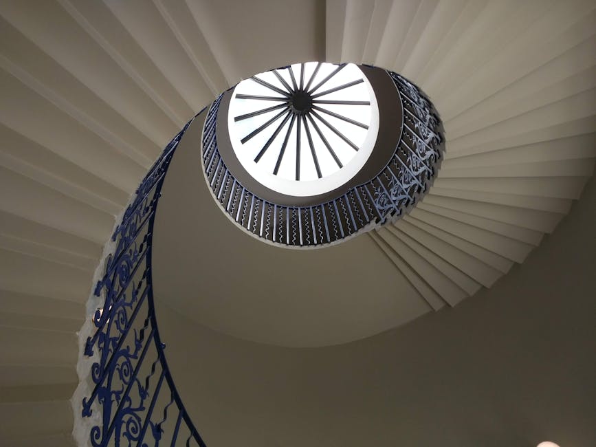 The image shows a spiral staircase viewed from below, with white steps and a decorative dark blue wrought iron railing featuring intricate scrollwork. Natural light illuminates the staircase from a circular skylight at the top, creating a bright central opening. The staircase curves upward in a helical shape within an interior space, with the smooth, light-colored wall providing a neutral backdrop. The upward perspective highlights the architectural detail of the staircase, which is likely part of a historic or elegant property, relevant to house removals and home relocation services offered by Removal Companies Greenwich, particularly in contexts involving packing, furniture transport, or moving logistics near the Cutty Sark area.