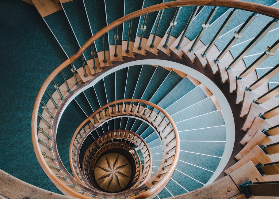 A top-down view of a spiral staircase inside a building, featuring wooden handrails and metal balusters with round finials supporting a continuous wooden handrail. The staircase has dark blue carpeted treads with white risers, arranged in a precise spiral pattern, leading downward towards a light wooden floor with a decorative star-shaped pattern at the base. The surrounding walls are painted in a neutral shade, and natural lighting illuminates the scene, highlighting the elegant curves and the smooth finish of the wood. This image illustrates the interior environment where a home relocation or moving service, such as those offered by Removal Companies Greenwich, might involve navigating staircases during furniture transport or packing and moving procedures.