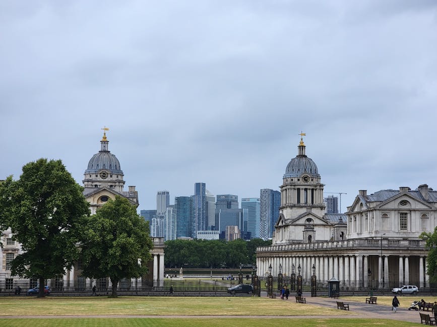 A wide view of central Greenwich with historic Christopher Wren-designed church buildings featuring domed roofs and classical columns situated on a grassy area. In the background, modern high-rise office buildings with glass facades rise into a cloudy sky, indicating an urban environment. In the foreground, there are several cardboard boxes, some wrapped in plastic film, and furniture pieces such as wooden chairs and tables arranged near the entrance of the churches. A trolley and moving blankets are visible, used for furniture transport or packing during home relocation. The scene shows a busy moving process, with people walking and loading items into a van parked nearby, illustrating the logistical aspects of removals and packing in Greenwich. Removal Companies Greenwich manage such home and furniture moving activities, coordinating loading and unloading in city and residential areas.