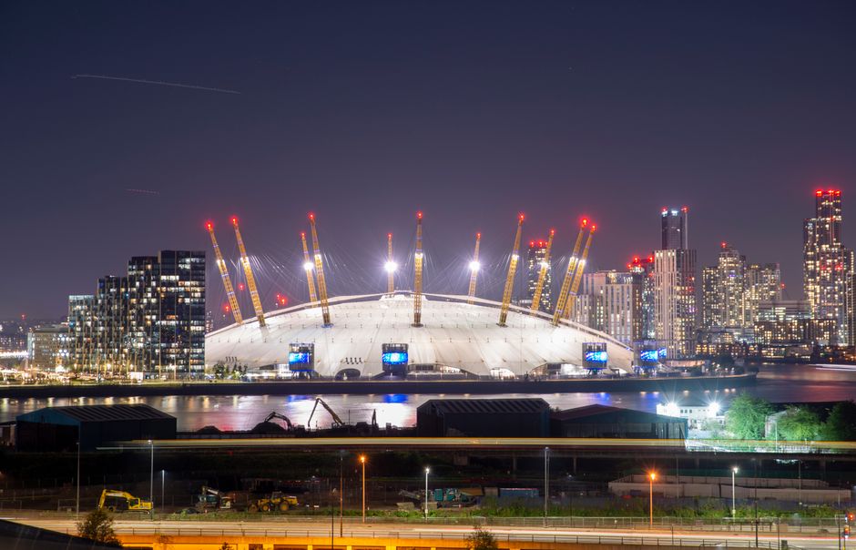 A wide view of central Greenwich with historic Christopher Wren-designed church buildings featuring domed roofs and classical columns situated on a grassy area. In the background, modern high-rise office buildings with glass facades rise into a cloudy sky, indicating an urban environment. In the foreground, there are several cardboard boxes, some wrapped in plastic film, and furniture pieces such as wooden chairs and tables arranged near the entrance of the churches. A trolley and moving blankets are visible, used for furniture transport or packing during home relocation. The scene shows a busy moving process, with people walking and loading items into a van parked nearby, illustrating the logistical aspects of removals and packing in Greenwich. Removal Companies Greenwich manage such home and furniture moving activities, coordinating loading and unloading in city and residential areas.