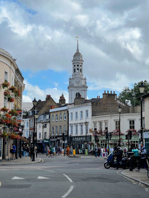 A busy street scene in Greenwich with pedestrians walking on the pavement and crossing the road, some carrying shopping bags or waiting at crosswalks. Several shops with shopfronts and signage line the street, including a café and retail stores. In the foreground, a delivery scooter is parked near the curb, while other vehicles, including cars and vans, are visible on the road. The background features historic buildings with traditional façades, and a prominent white clock tower with a spire rises above the rooftops, set against a partly cloudy sky. This setting illustrates the vibrant urban environment typical of Greenwich, where house removals and packing services by Removal Companies Greenwich could be coordinated for moving or transport logistics within the area.