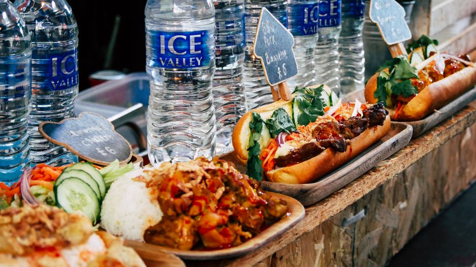 Inside a market stall, several trays of food are displayed on a wooden counter, including plates of curry with rice, fresh sliced cucumber and carrot garnishes, and sandwiches filled with ingredients like grilled meat, vegetables, and herbs, all arranged for serving or takeaway. Behind the food, there are multiple large bottles of branded 'Ice Valley' bottled water, positioned in a row along the counter. Small chalkboard signs with handwritten prices are placed next to some items. The scene is well-lit, highlighting the vibrant colours and textures of the food, with a focus on the presentation typical of market catering or quick-service food items. The setting suggests a typical scene of a market trader or caterer preparing for or during a home relocation or market trading activity, with Removal Companies Greenwich providing logistics and packing support for market traders.