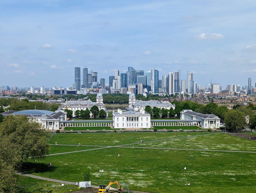 A wide-angle view of a historic white palace with classical architecture, situated on a well-maintained green lawn with pathways and scattered visitors. The palace features symmetrical wings, tall columns, and turreted rooftops, with some windows open. In the foreground, people are walking, sitting, and relaxing on the grass, while a few are engaged in activities such as taking photos or strolling along the pathways. The scene appears to be taken during daylight on a partly cloudy day, with soft natural light illuminating the building and the surrounding park. In the background, a modern city skyline with numerous high-rise office buildings and skyscrapers dominates the horizon, contrasting sharply with the classical appearance of the palace. This setting captures the blend of historic and contemporary environments, suitable for describing house removals, furniture transport, and packing and moving processes managed by Removal Companies Greenwich during a same-day relocation from Maze Hill to Greenwich Park.