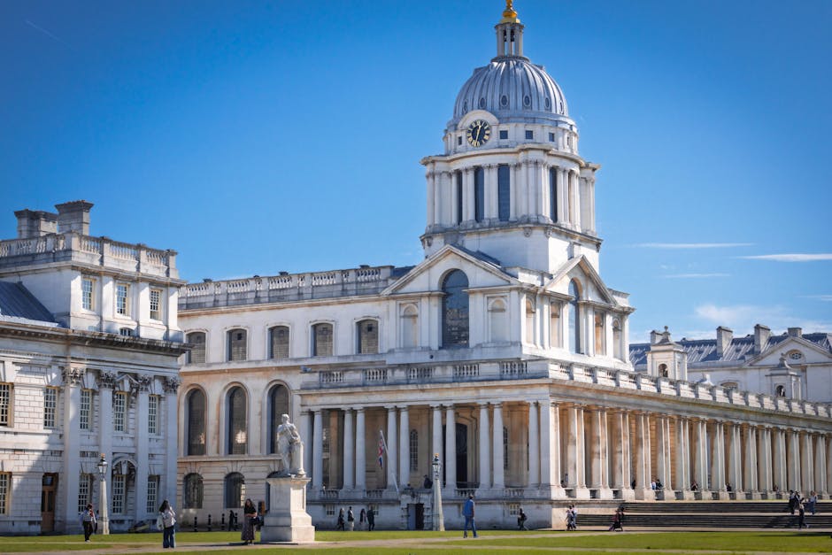 A large, historic white stone building with a prominent domed tower featuring a clock face, situated under a clear blue sky. The structure has classical architectural details, including a colonnade al