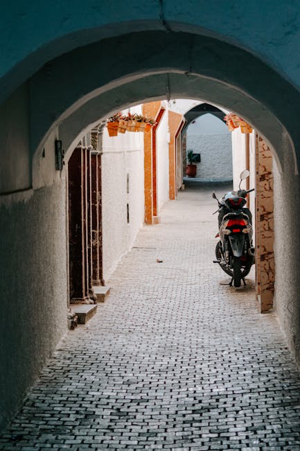 A narrow cobbled alleyway viewed through a rounded archway, with buildings on either side painted in light colors. Inside the alley, a black and red scooter is parked near a doorway, and behind it, a white door is open, revealing an interior space. Potted plants and hanging flower baskets decorate the area, and a potted plant is visible at the end of the alley. The scene appears to be part of a residential area, likely during daytime with natural lighting. This setting exemplifies a typical narrow street in Greenwich, suitable for house removals and furniture transport, with clean paving and organized curbside parking. The image is relevant for illustrating local moving logistics, highlighting the need for careful navigation through tight spaces during home relocation, aligning with the services offered by Removal Companies Greenwich in their SE10 house removals guide for narrow streets.