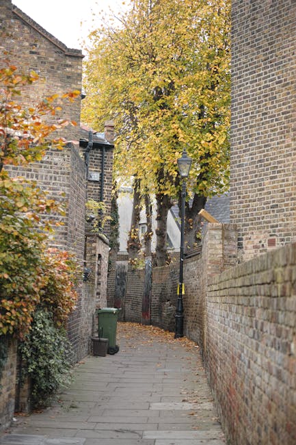 A narrow residential street in Greenwich with a steep incline, flanked by white and grey terraced houses on either side. On the left, there are small potted plants with green foliage placed along the pavement near the building wall. The right side features a dark grey door set into the white façade of a house. The pavement is made of asphalt with concrete edging, and there are visible drainage grates along the edge. In the distance, a larger white building with multiple windows is visible at the top of the hill, with overcast sky overhead. Beneath the image, there is no visible activity or moving equipment, but this setting is typical for a home relocation or furniture transport operation in the greenery-lined streets of Greenwich, where careful navigation of narrow streets is essential for successful removals. Removal Companies Greenwich may utilize such environments when providing house removals services specific to tightly packed urban streets like those in SE10.