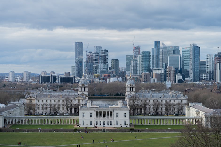 A wide-angle view of a large, historic white building with classical architecture, including columns, domed towers, and ornate details, situated in a spacious green park with pathways and scattered visitors. In the background, a modern city skyline features numerous high-rise office buildings, skyscrapers, and construction cranes under a cloudy sky. The scene captures the transition from historic architecture to contemporary urban development, with the greenery providing a buffer between the two environments. This setting illustrates a location suitable for house removals or home relocation services, with a focus on urban transport logistics and the moving process near prominent city landmarks, as provided by Removal Companies Greenwich.