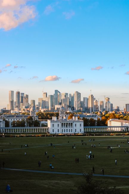 A wide view of a city skyline featuring modern glass skyscrapers and office buildings under a partly cloudy sky during daylight. In the foreground, a large open park area with well-maintained grass hosts groups of people walking, sitting, and engaging in recreational activities, with some individuals children playing on the grass. There is a white, single-story building near the park's edge, possibly a pavilion or small facility, with a pathway surrounding it. The scene is illuminated by natural light, emphasizing the contrast between the urban high-rise structures in the background and the greenery of the park in the foreground. This image captures the urban landscape typical of a busy city centre, relevant to themes of residential relocations and moving logistics supported by companies like Removal Companies Greenwich.