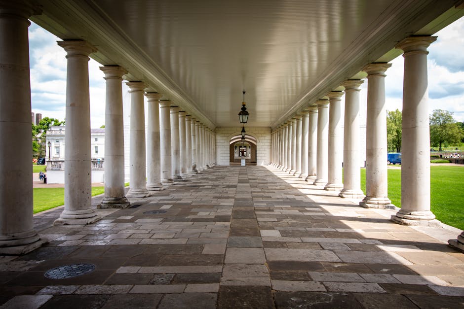 A wide-angle view of a large, historic white building with classical architecture, including columns, domed towers, and ornate details, situated in a spacious green park with pathways and scattered visitors. In the background, a modern city skyline features numerous high-rise office buildings, skyscrapers, and construction cranes under a cloudy sky. The scene captures the transition from historic architecture to contemporary urban development, with the greenery providing a buffer between the two environments. This setting illustrates a location suitable for house removals or home relocation services, with a focus on urban transport logistics and the moving process near prominent city landmarks, as provided by Removal Companies Greenwich.